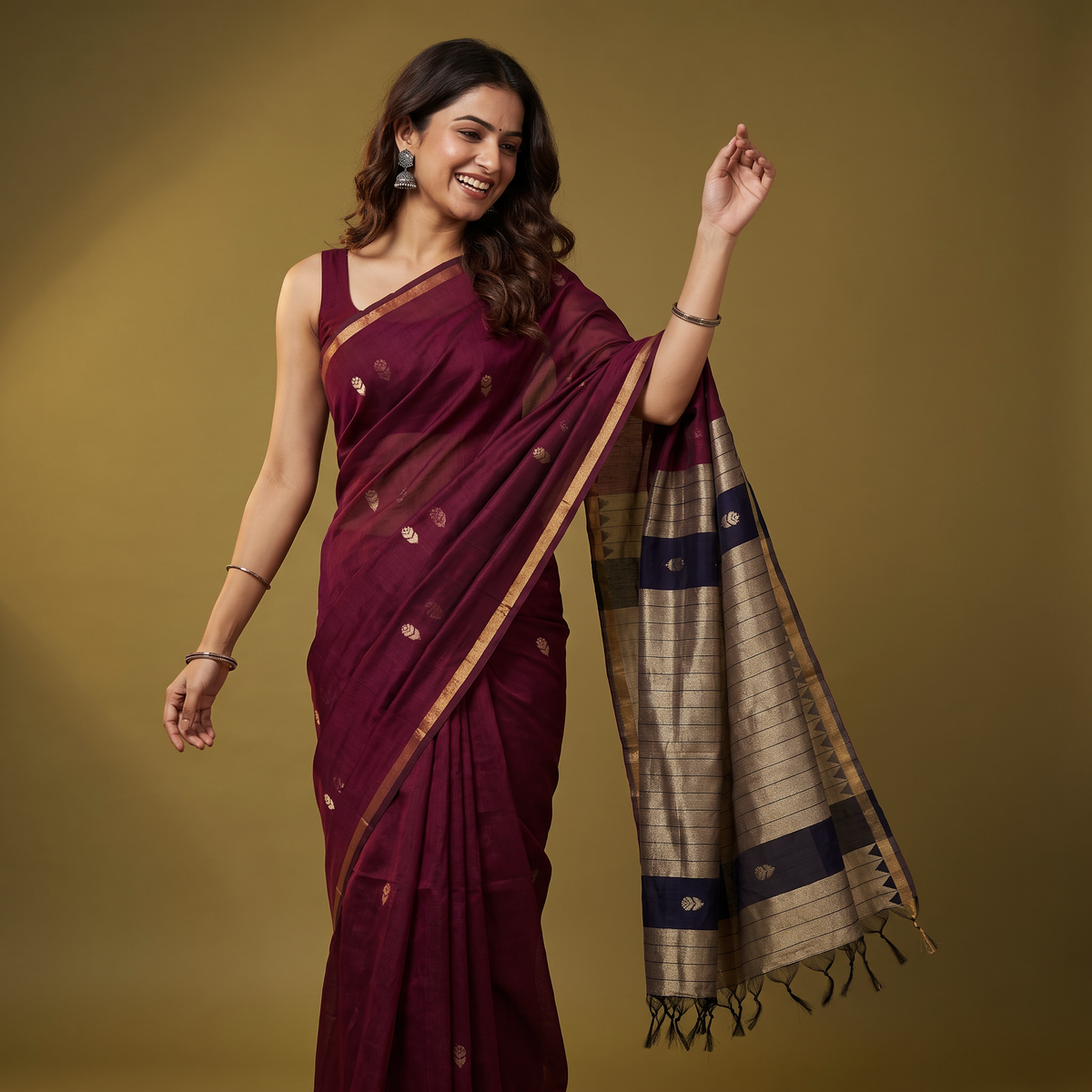A woman modeling a deep maroon silk saree with gold zari motifs and a beige and navy blue patterned pallu, posed against a solid olive-brown studio background.