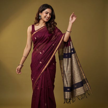 A woman modeling a deep maroon silk saree with gold zari motifs and a beige and navy blue patterned pallu, posed against a solid olive-brown studio background.